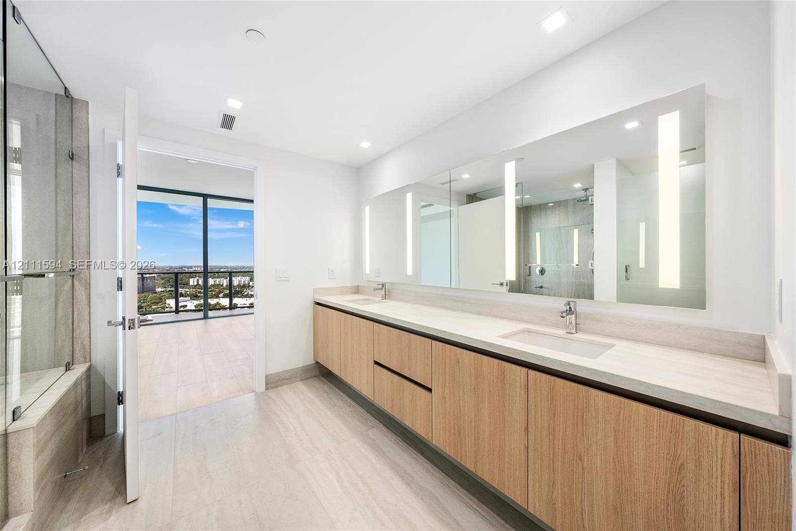 2411 Laguna Circle, #2506, North Miami, FL 33181 Floating vanity with wood-finish cabinetry, dual under-mount sinks, expansive wall-to-wall integrated mirror with vertical task lighting, and glass-enclosed shower area