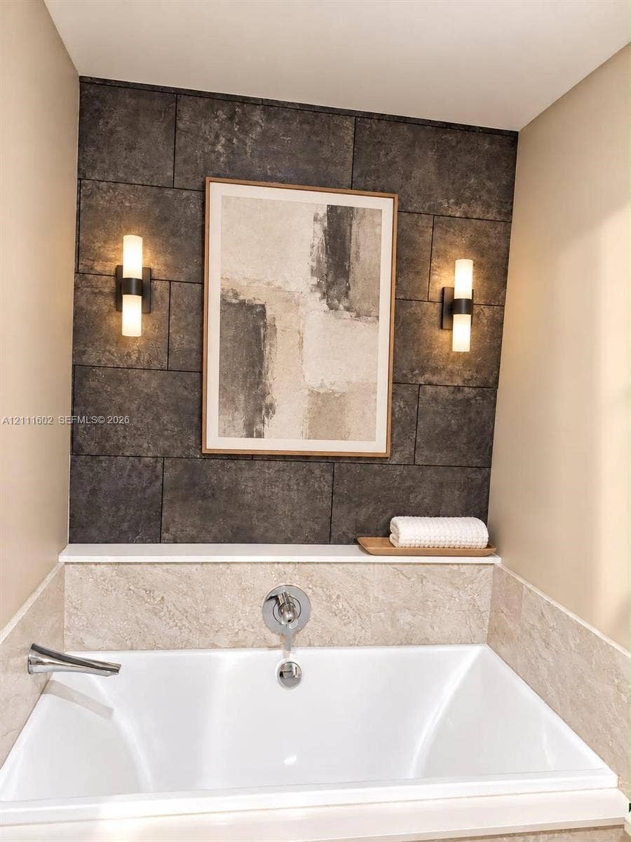 Built-in soaking tub featuring a neutral-toned stone tile surround, a dark stone tile accent wall, integrated display ledge, and dual wall-mounted cylindrical light fixtures