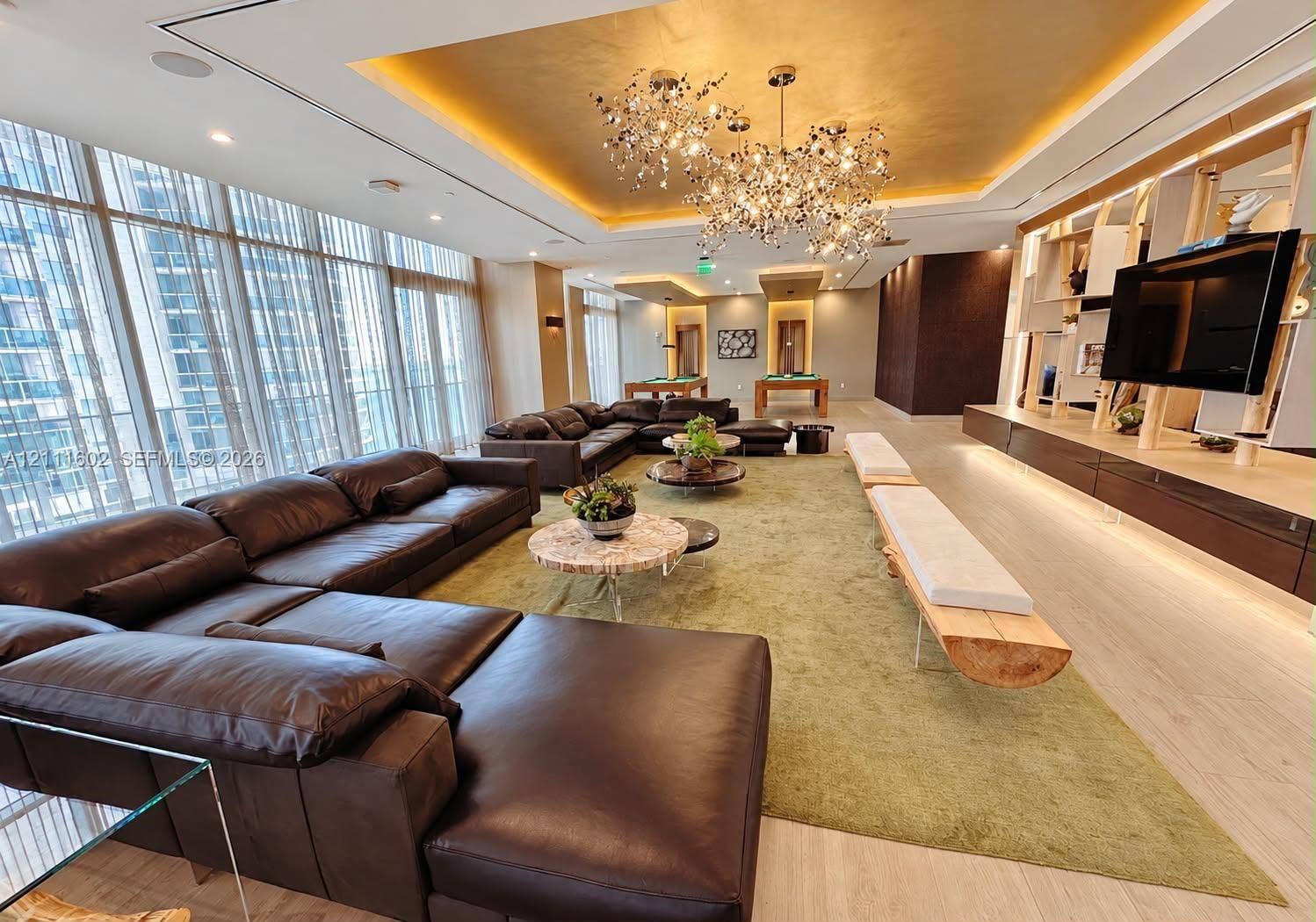 Expansive room featuring floor-to-ceiling windows, gold-leaf tray ceiling, branching metallic statement chandelier, light-toned wood-finish flooring, and extensive custom built-in cabinetry with integrated media shelving