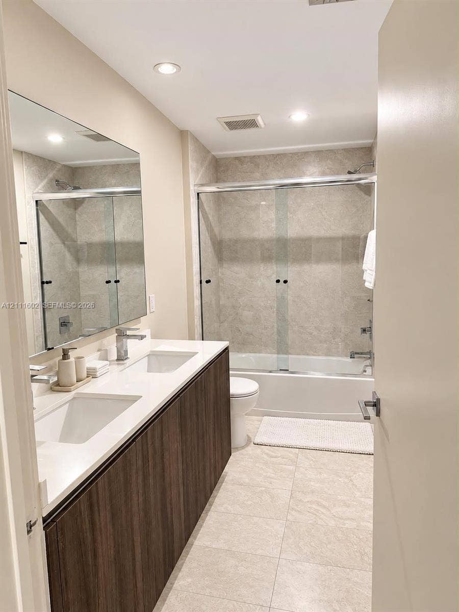 Dual-sink vanity with wood-finish cabinetry, quartz countertop, large wall-mounted mirror, and tub-shower combination featuring sliding glass doors and neutral tile surround