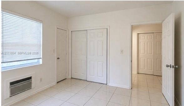 Interior space featuring ceramic tile flooring, neutral walls, two sets of six-panel closet doors, a multi-pane window with adjustable blinds, and a built-in through-wall air conditioning unit