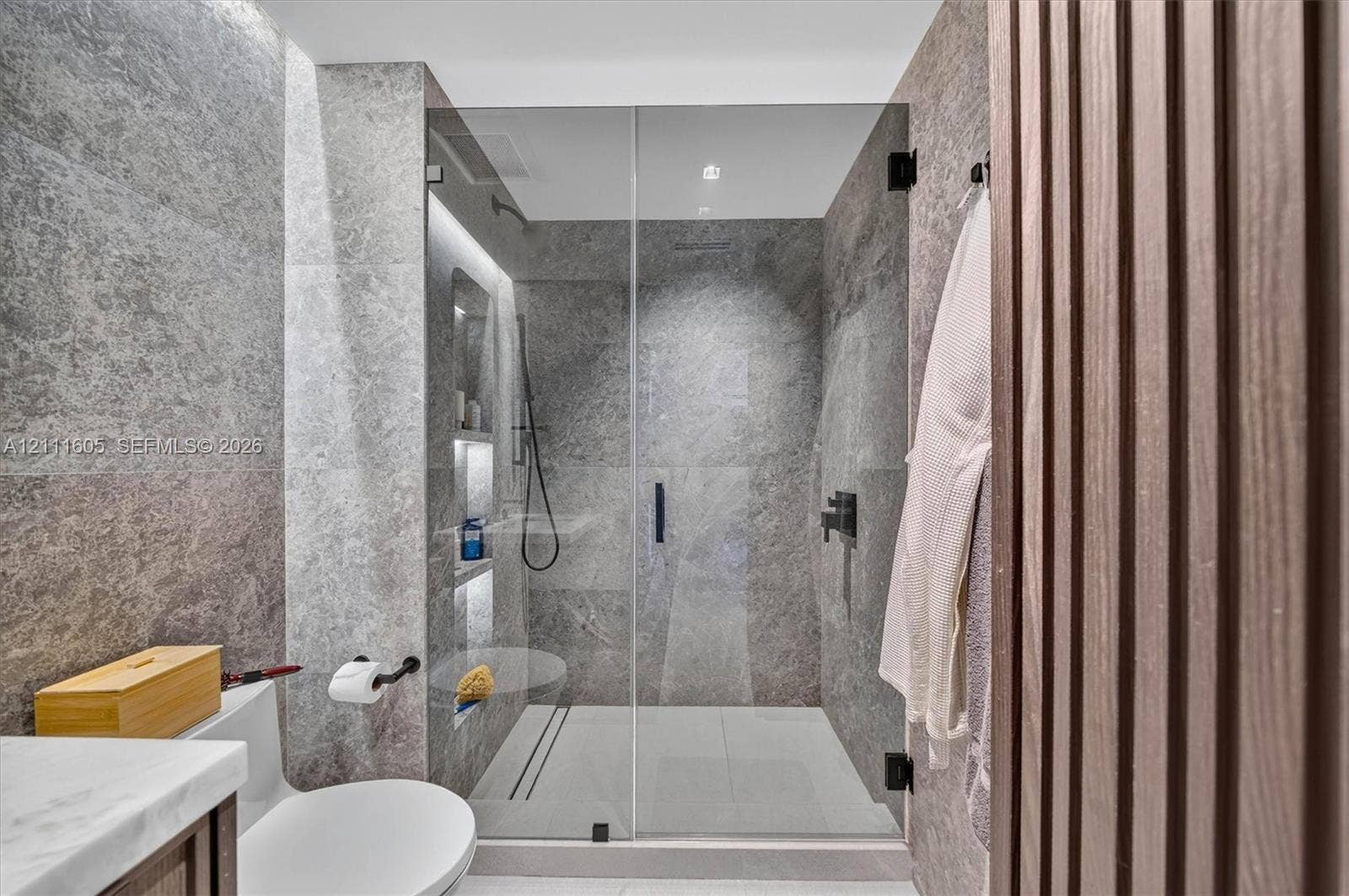 Walk-in shower with frameless glass enclosure, matte black fixtures, integrated wall shelving, floor-to-ceiling stone tile, and vertical wood-finish paneling
