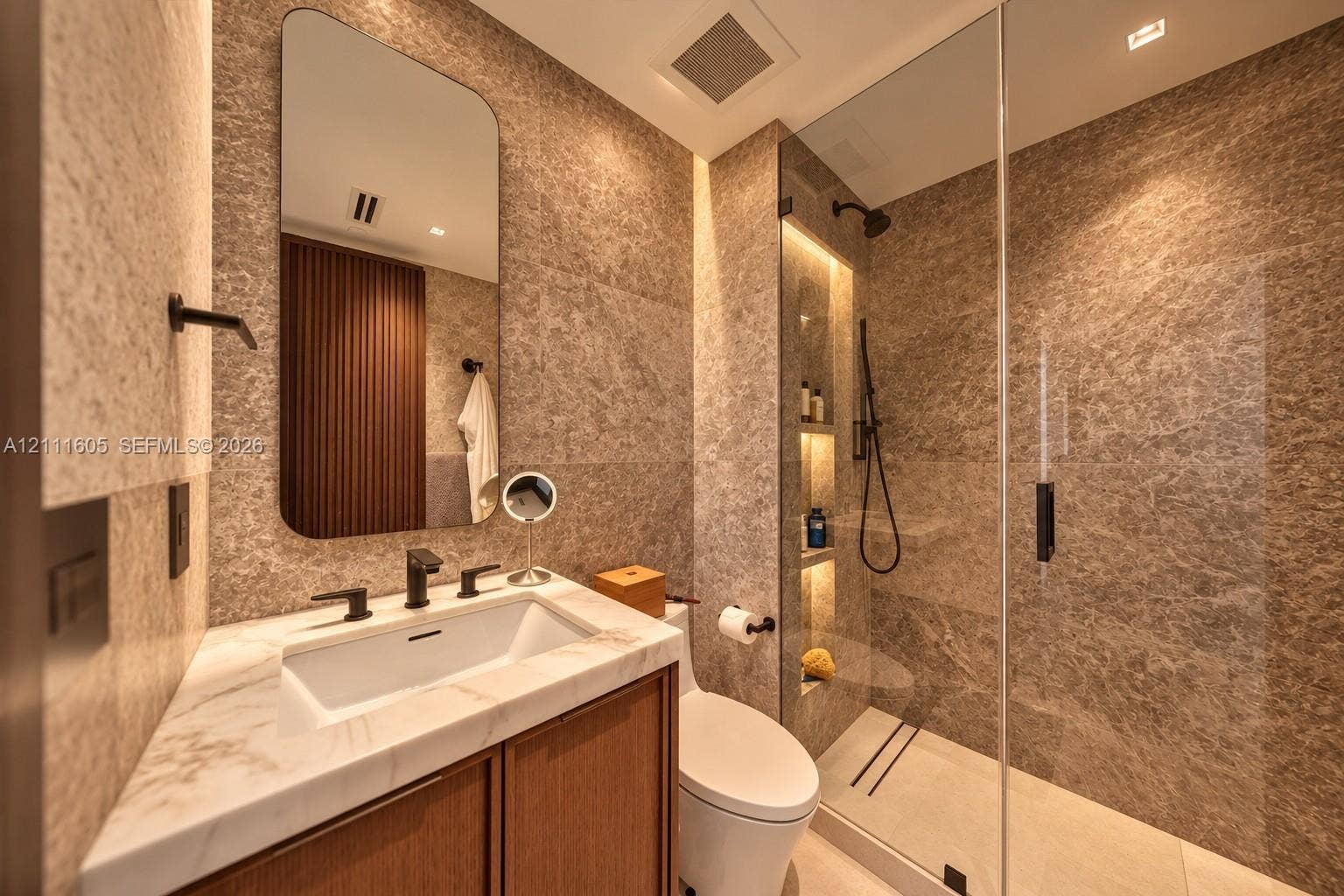Bathroom featuring floor-to-ceiling stone tile, glass-enclosed shower with integrated recessed shelving, wood-finish vanity cabinet, stone countertop with undermount sink, and matte black fixtures