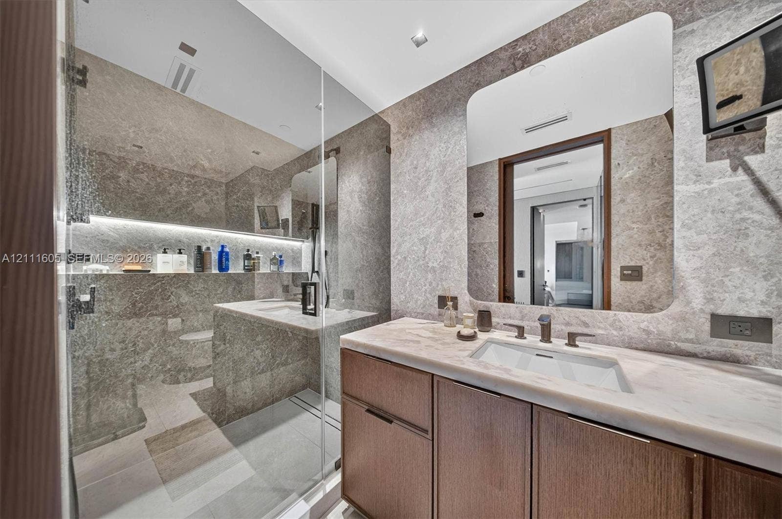 Custom wood-finish vanity with integrated stone countertop, undermount sink, stone tile wall surfaces, and frameless glass shower enclosure