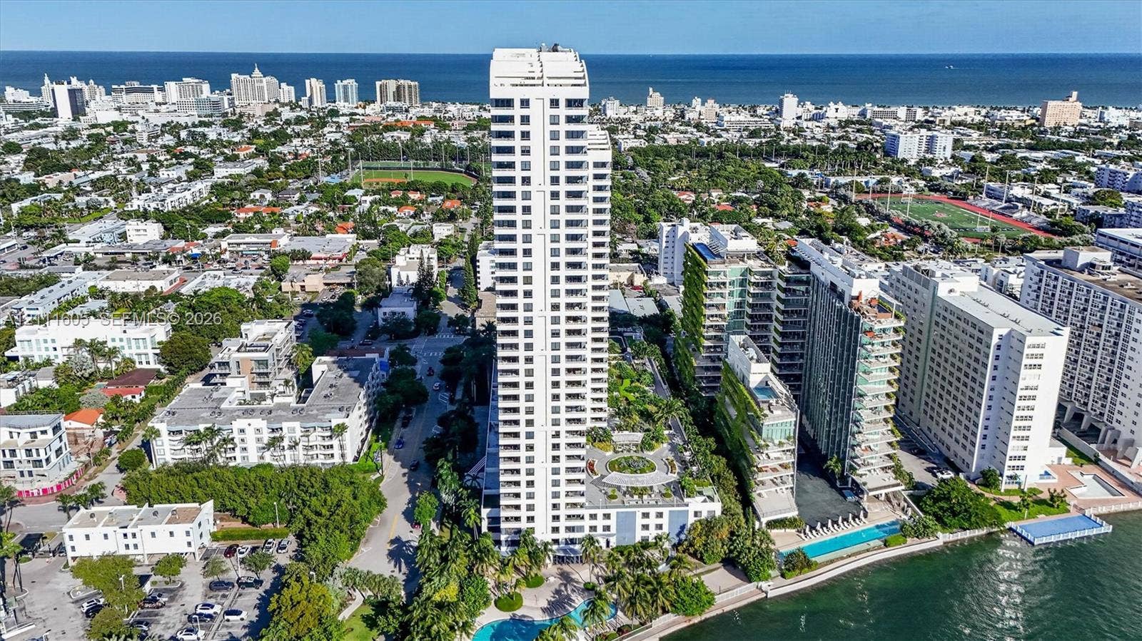 High-rise residential tower featuring a white facade, landscaped podium-level terrace, ground-level swimming pool, and water-front placement