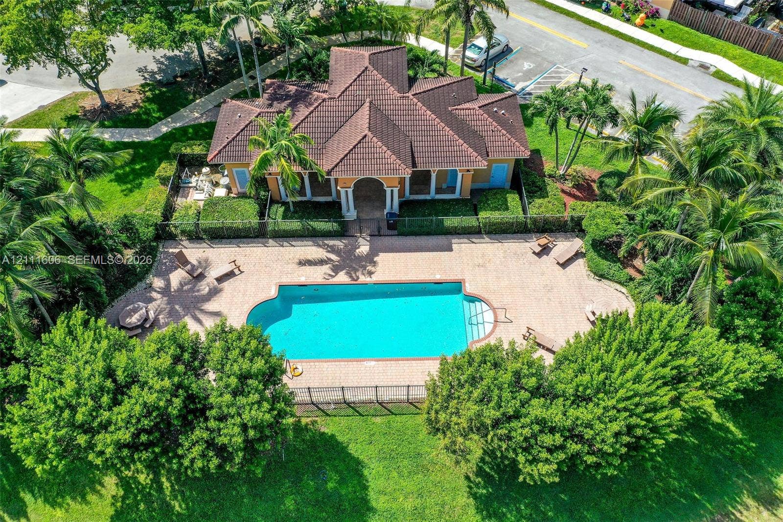 Single-story building featuring a tile roof, an expansive stone-paver pool deck, a swimming pool with built-in stairs, perimeter fencing, and mature palm trees