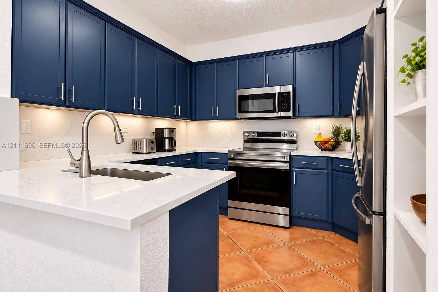 Kitchen featuring navy blue cabinetry, stainless steel appliances, light-toned stone-finish countertops, under-cabinet lighting, and ceramic tile flooring