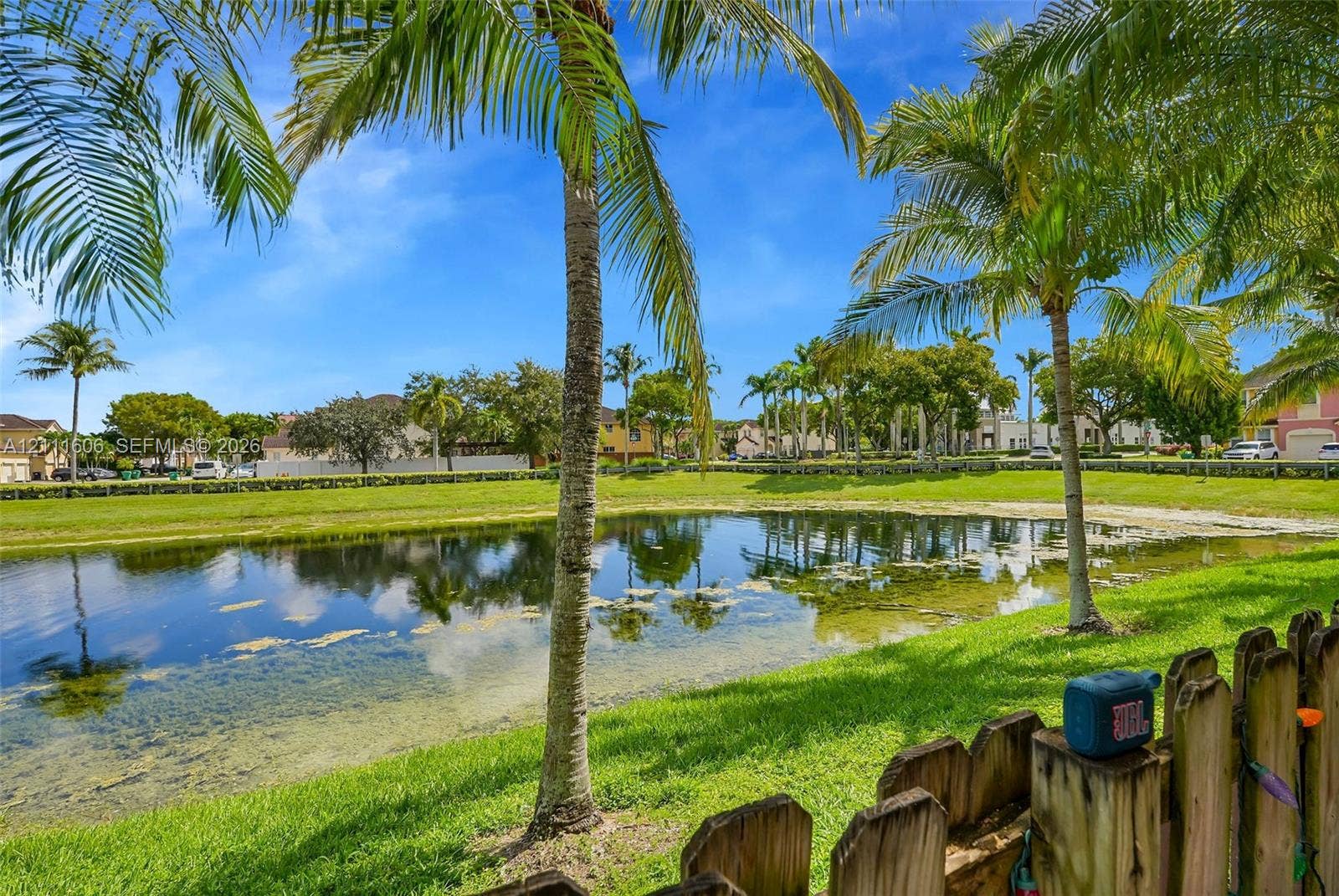 Waterfront lot featuring a pond, manicured lawn, and mature palm trees enclosed by a wooden picket fence