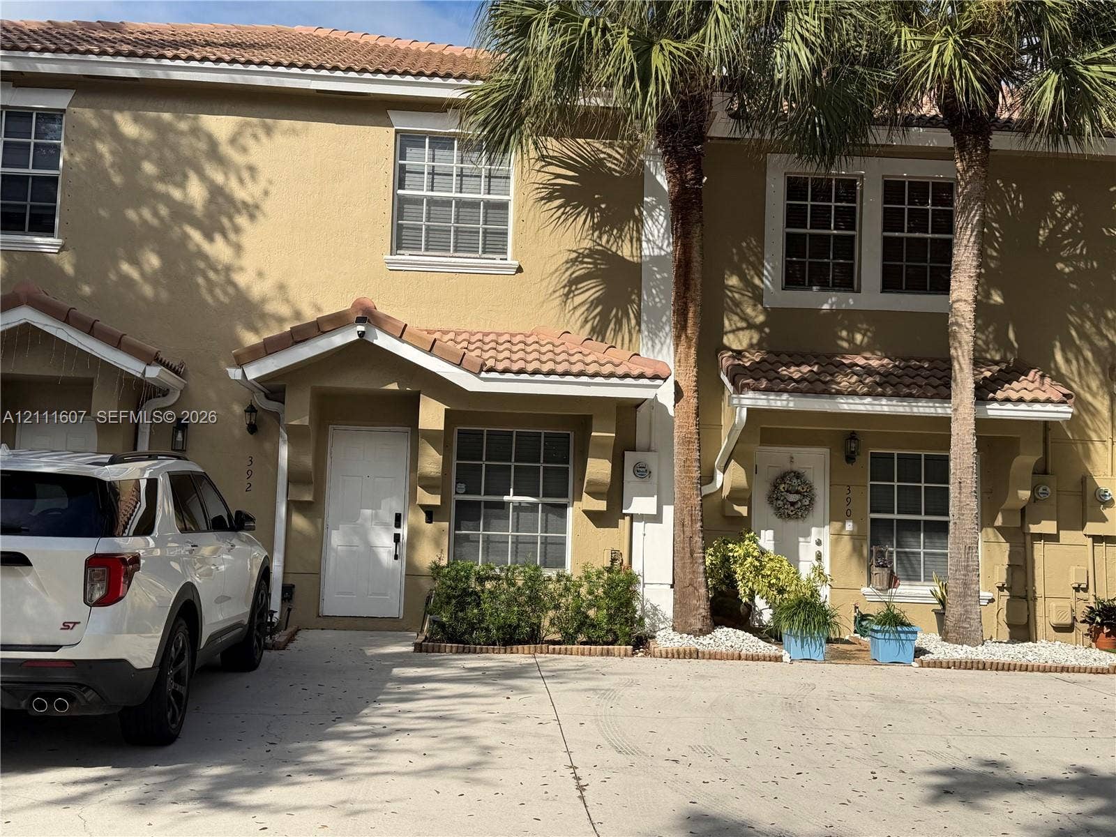 Two-story townhome exterior featuring a stucco facade, tile roof, gabled entry overhangs, and mature palm trees