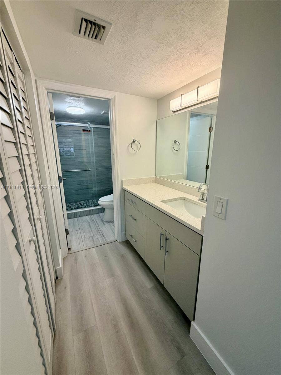 Bathroom featuring a vanity with stone-finish countertop, undermount sink, linear wall-mounted lighting, wood-finish flooring, and a glass-enclosed shower