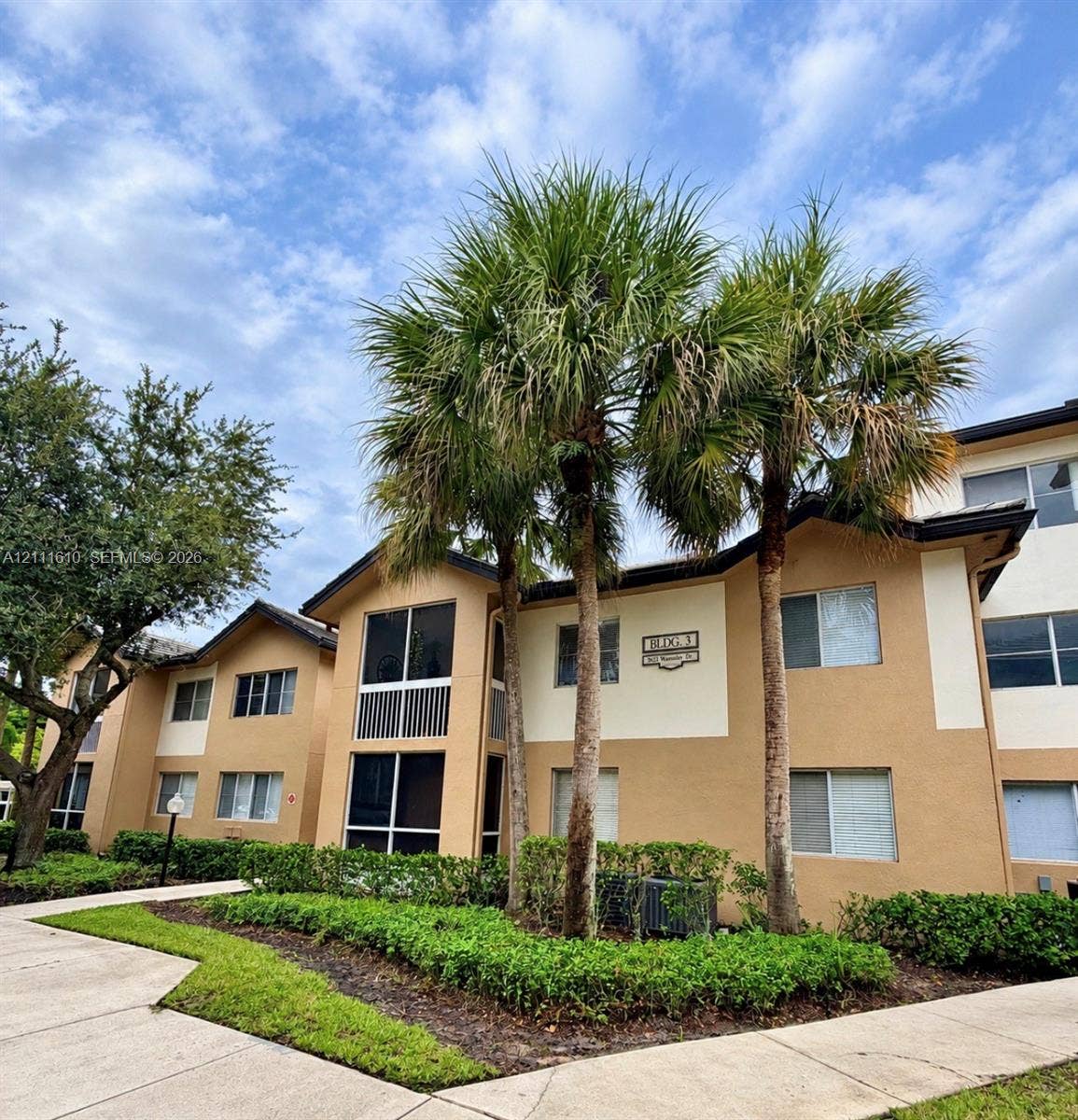 Multi-story exterior featuring a neutral stucco facade, screened-in balconies, mature palm trees, manicured hedging, and concrete walkways