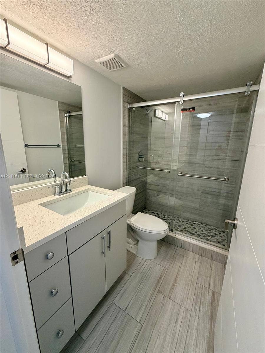 Bathroom featuring a quartz-topped vanity with an undermount sink, walk-in shower with a sliding glass door and pebble-stone floor, wood-finish floor tile, and wall-mounted vanity lighting