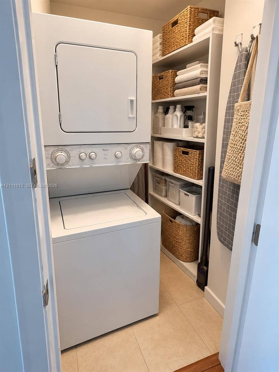 Laundry utility space featuring a stacked washer and dryer unit, built-in floor-to-ceiling white shelving, and neutral tile flooring