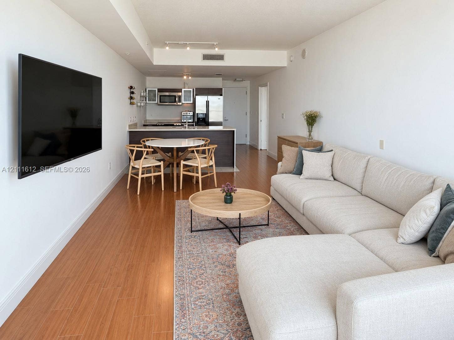 Open-concept floor plan featuring wood-finish flooring, recessed track lighting, and a kitchen equipped with dark cabinetry, stainless steel appliances, and a breakfast bar