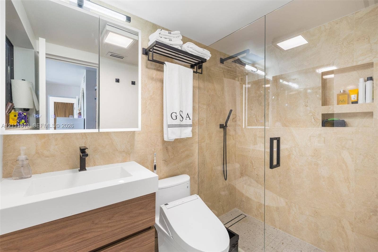 Bathroom featuring a wood-finish floating vanity with an integrated white sink, stone-tiled wall surfaces, a glass-enclosed shower with an integrated recessed shelf, and matte black hardware