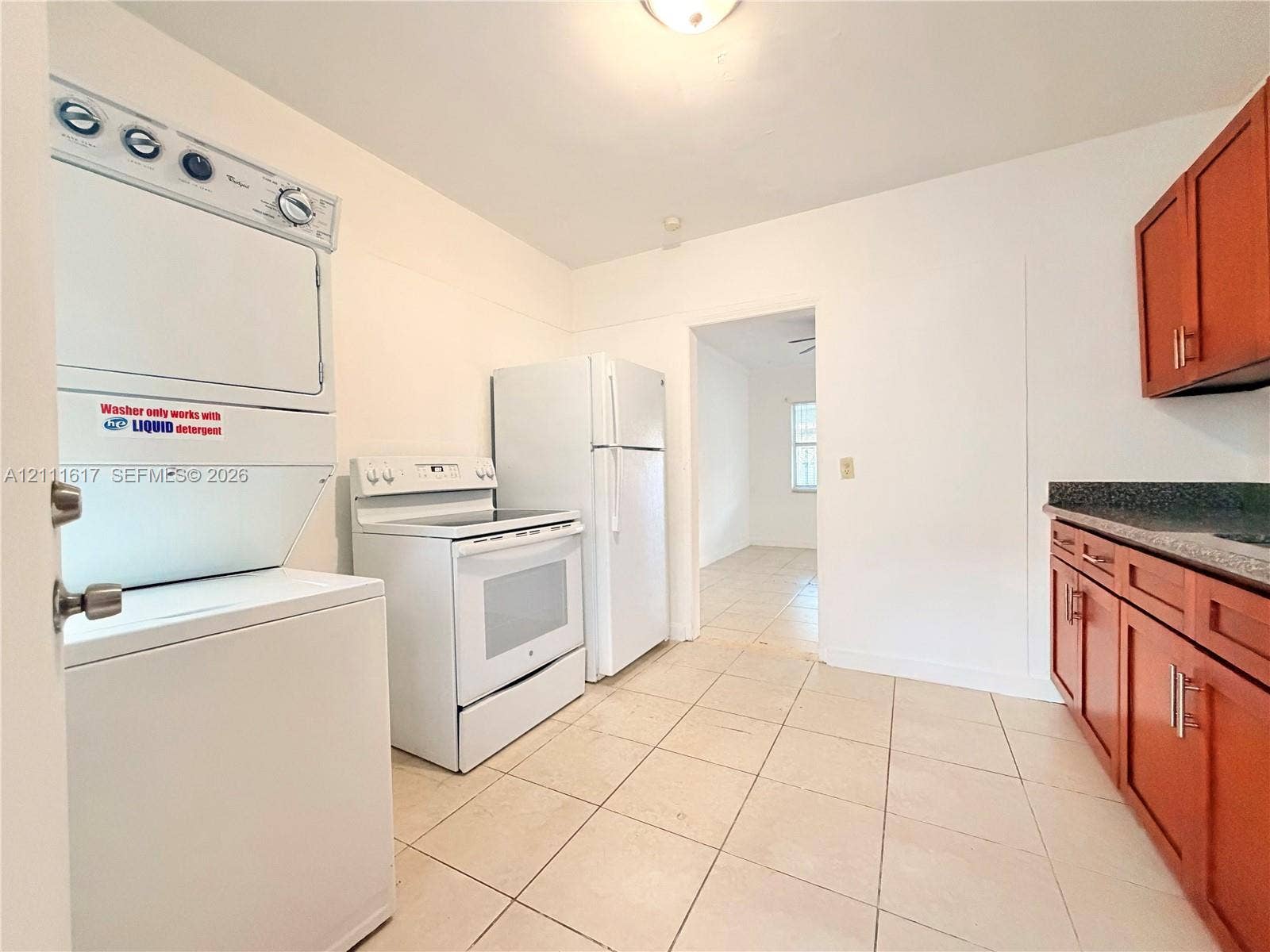 Kitchen featuring tile flooring, wood-finish cabinetry, dark solid-surface countertops, and a stacked laundry unit
