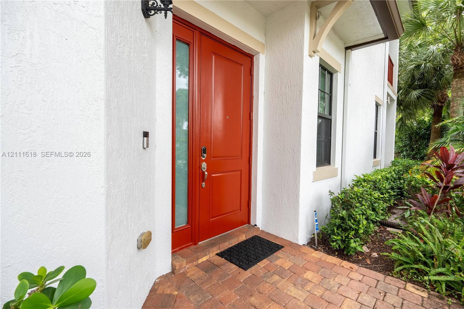 1423 SW 115th Avenue, #NA, Pembroke Pines, Pembroke Pines, FL 33025 Exterior entryway featuring a red solid-core front door with an integrated sidelight, stucco facade, brick paver flooring, and decorative corbel accents