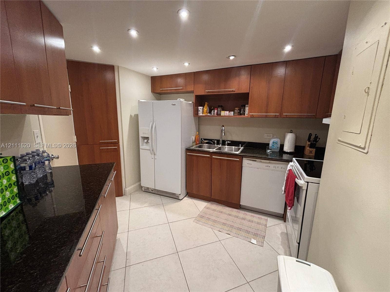 Kitchen featuring wood-finish cabinetry, dark stone-surfaced countertops, a double-basin stainless steel sink, recessed lighting, and large-format tile flooring