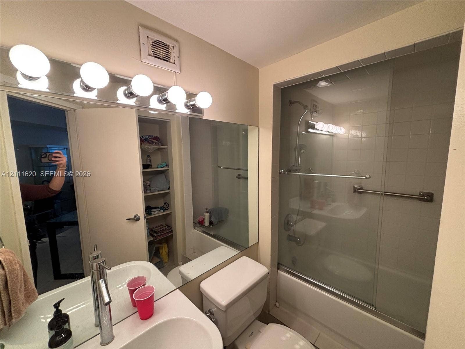 Bathroom featuring a single-sink vanity with a solid-surface countertop, a full-width wall mirror, a bathtub-shower combination with sliding glass doors and a grab bar, and built-in shelving
