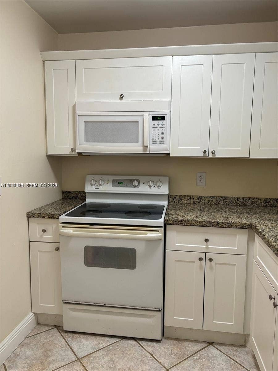 Kitchen featuring white shaker-style cabinetry, a stone-finish countertop, a built-in over-the-range microwave, a white electric range, and tile flooring