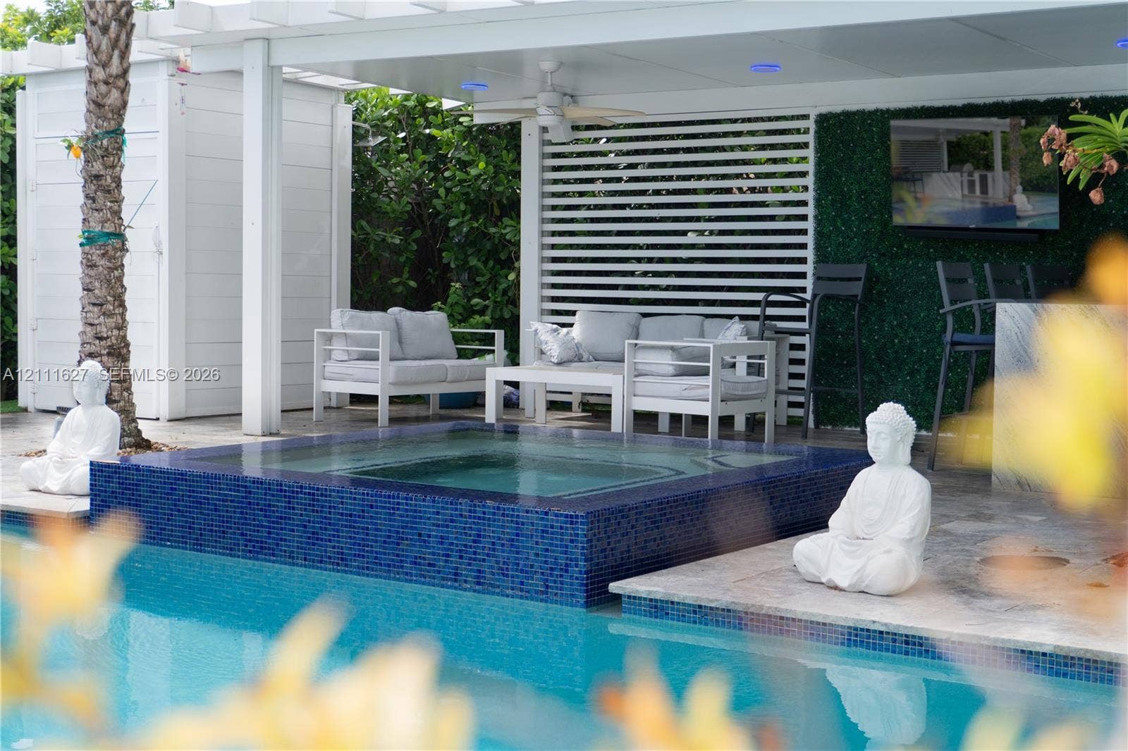 Covered outdoor lounge area featuring a blue mosaic tile spa, stone-finish decking, white slatted privacy screen, and recessed ceiling lighting