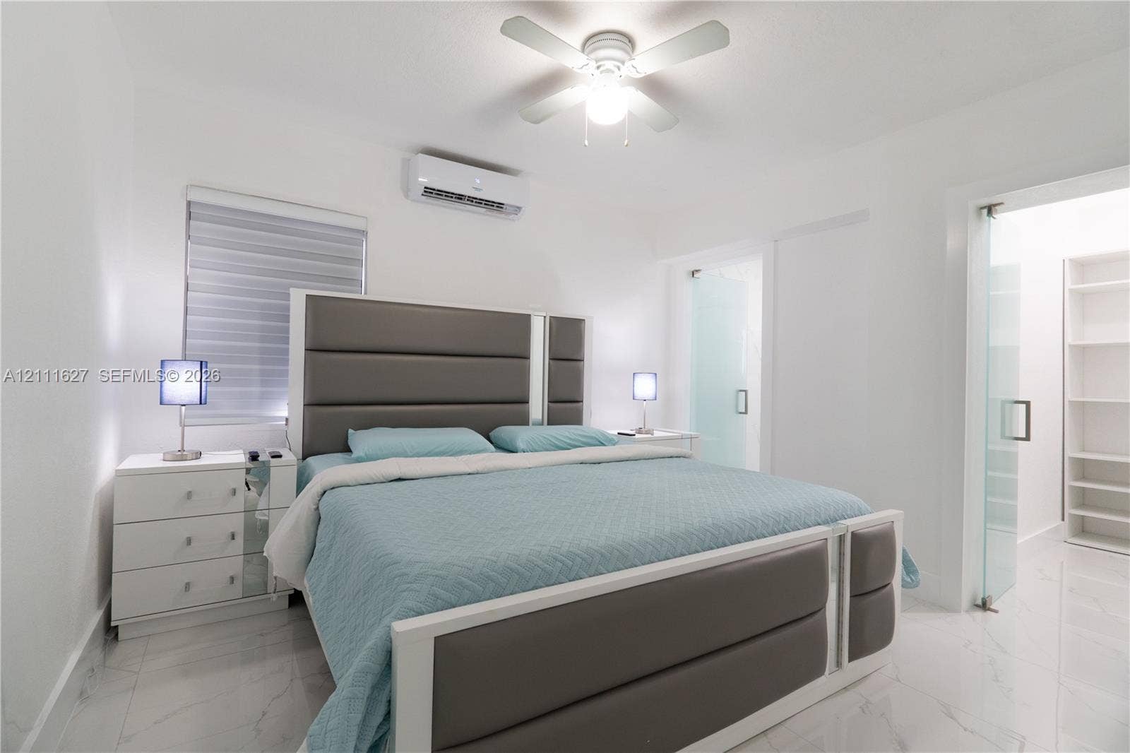 Neutral-toned interior featuring large-format tile flooring, a wall-mounted ductless air conditioning unit, a ceiling fan with integrated lighting, and a glass-paneled entry to a built-in shelving unit