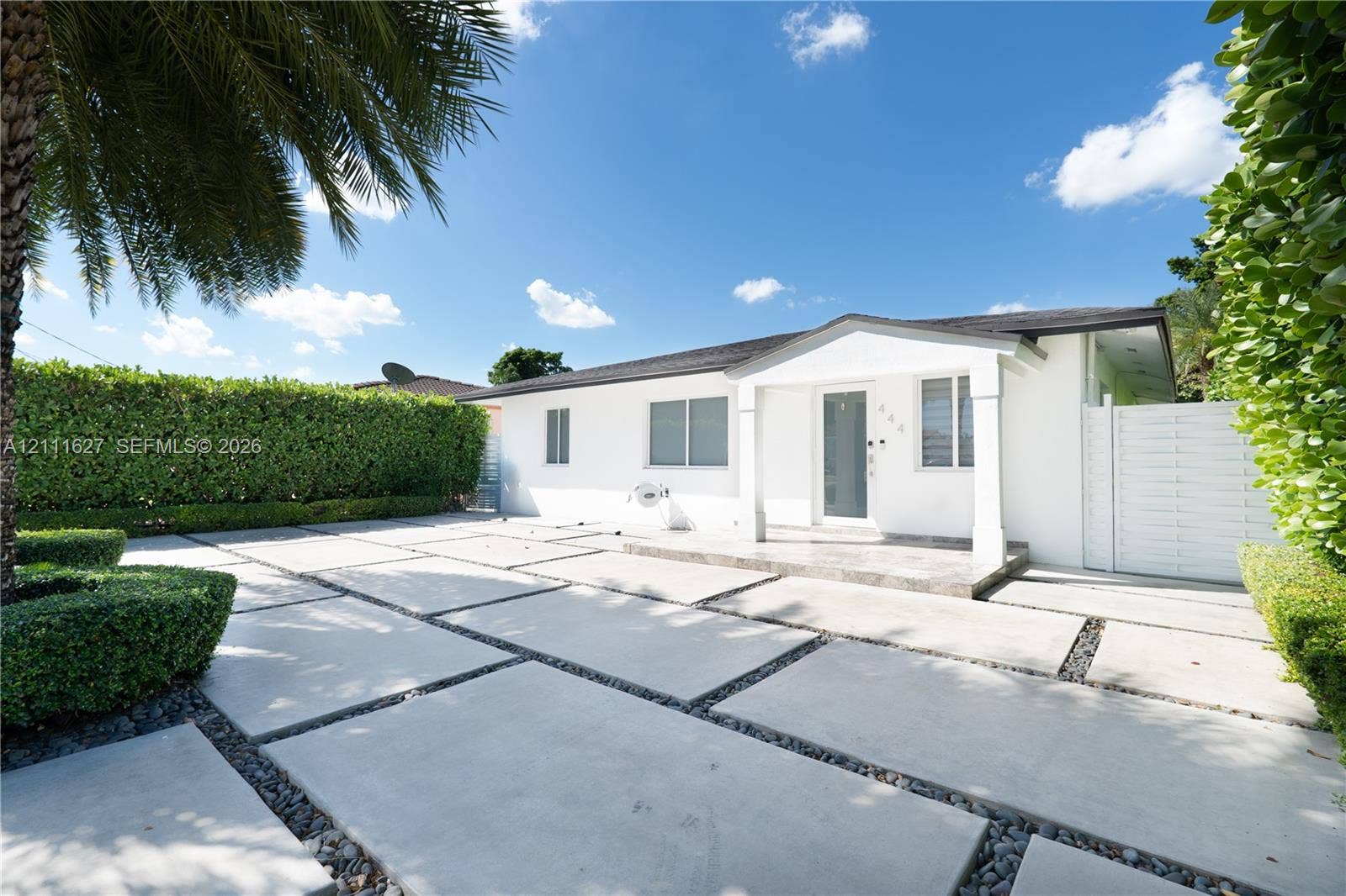 Exterior facade featuring a covered entryway, smooth white stucco finish, geometric concrete paver driveway with decorative stone accents, and manicured perimeter hedges