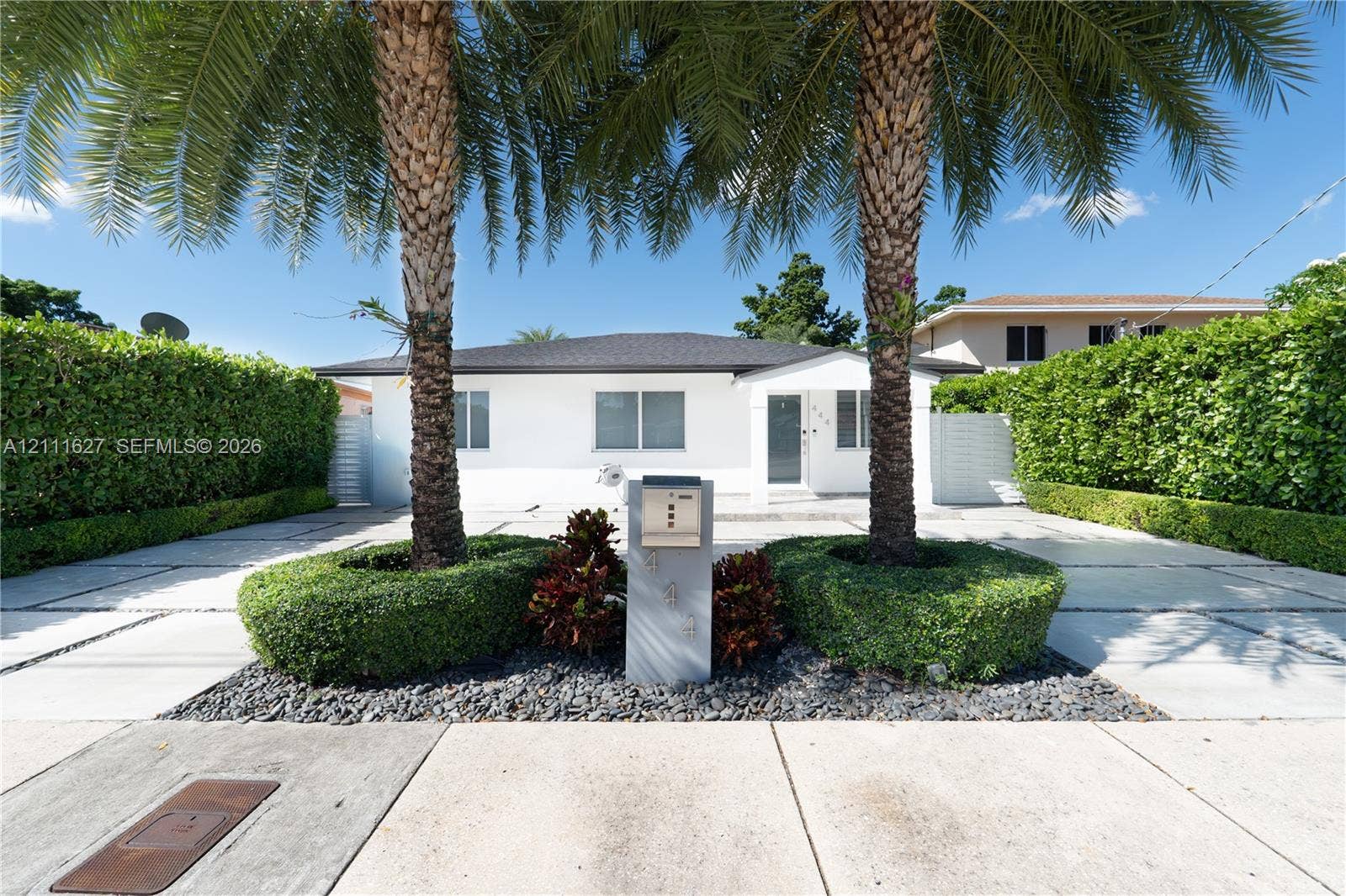 Single-story residence featuring a paved driveway with turf accents, mature palm trees, groomed privacy hedges, and a contemporary concrete mailbox pillar