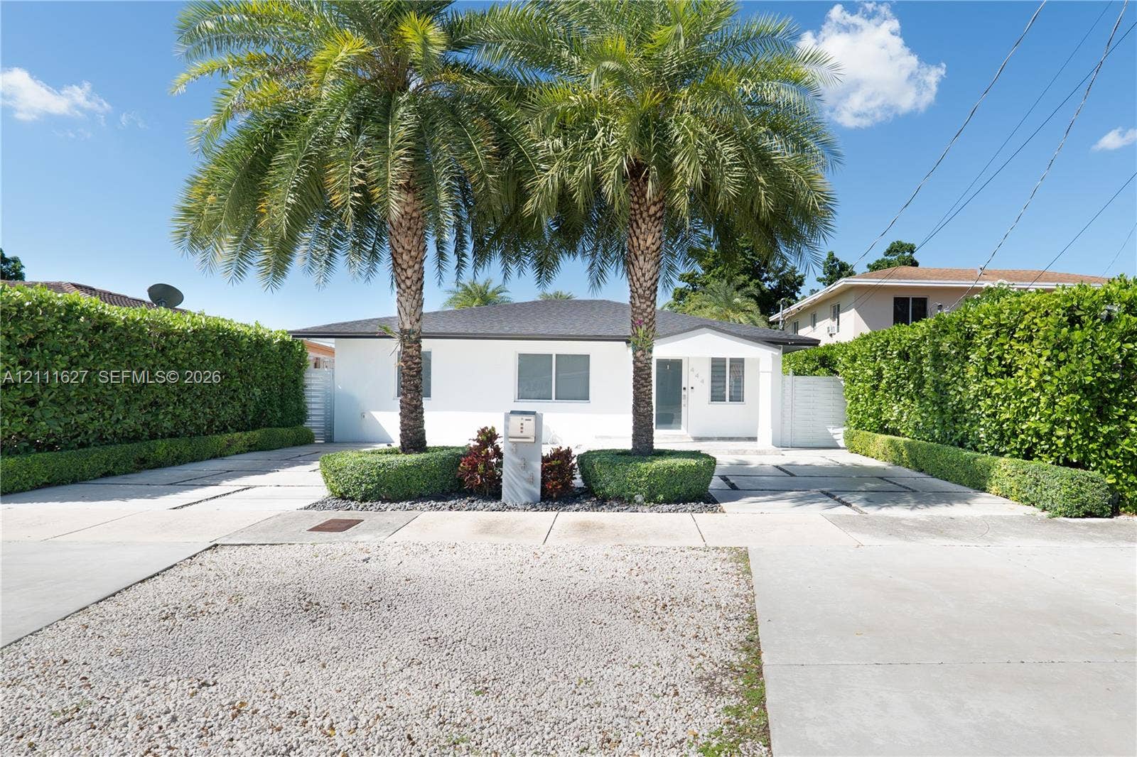 Single-story exterior featuring white stucco siding, a hip roof, expansive concrete paved driveway with integrated stone borders, mature palm trees, and dense perimeter hedge landscaping