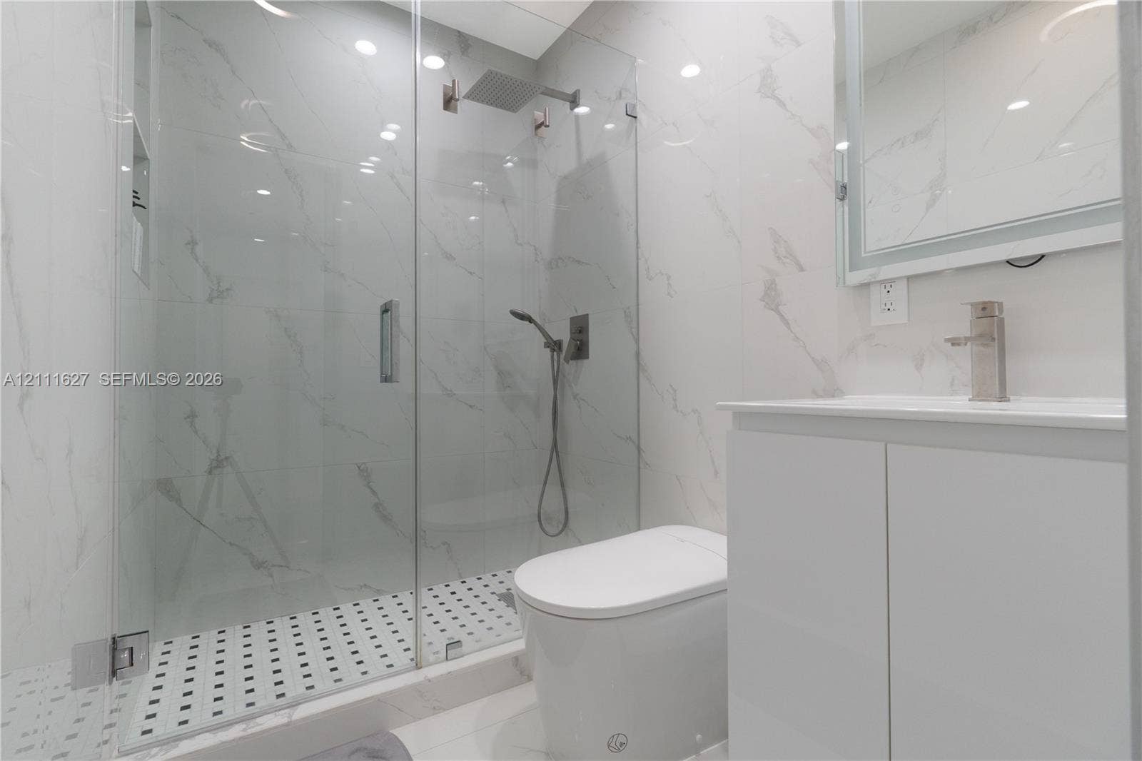 Walk-in glass-enclosed shower with a rainfall showerhead, large-format tile surround, mosaic tile flooring, and integrated wall niche, complemented by a white vanity with a metallic faucet and integrated lighting