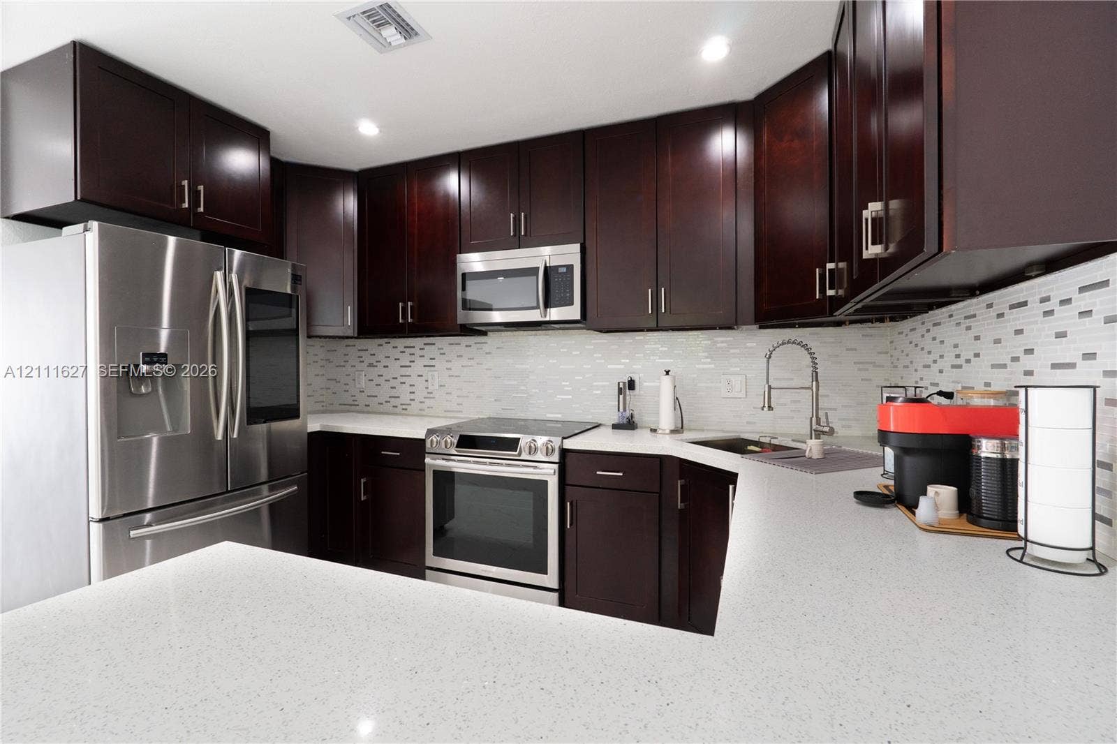 Kitchen featuring dark wood cabinetry, quartz countertops, a multi-tonal mosaic tile backsplash, and stainless steel appliances including a microwave, range, and refrigerator