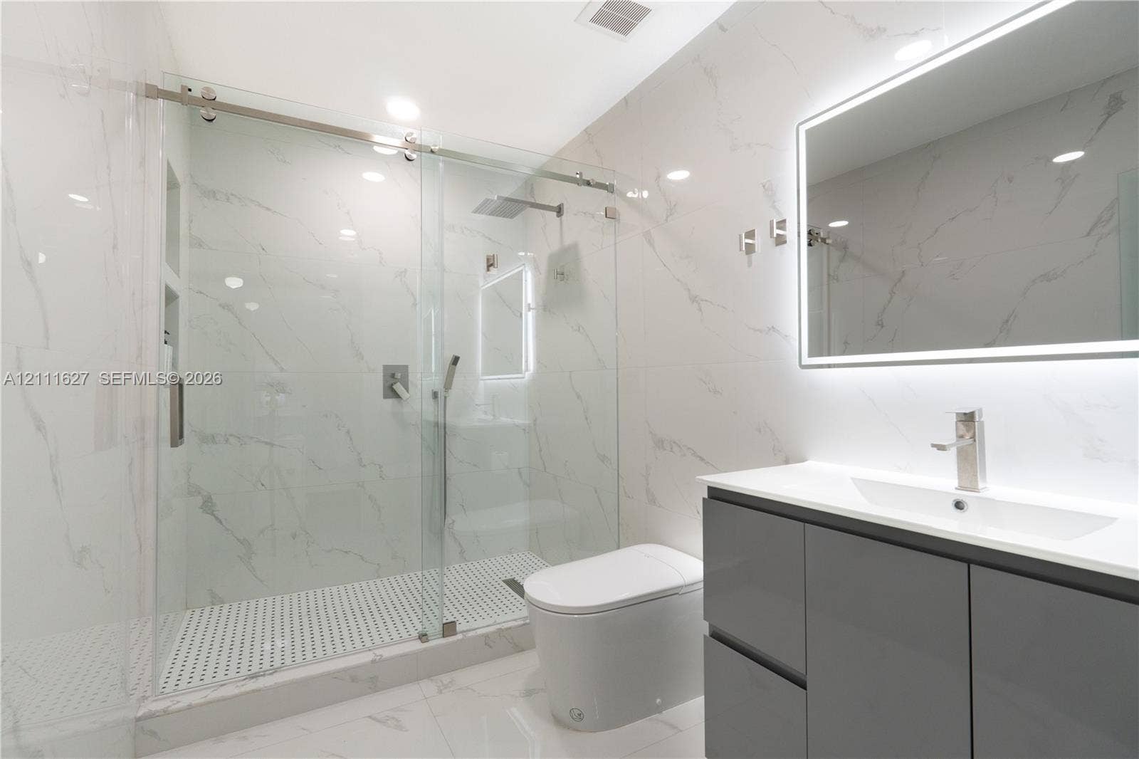 Walk-in glass-enclosed shower featuring stone-finish wall tile, rainfall showerhead, and built-in recessed shelving, complemented by a modern floating vanity with integrated lighting and a streamlined toilet