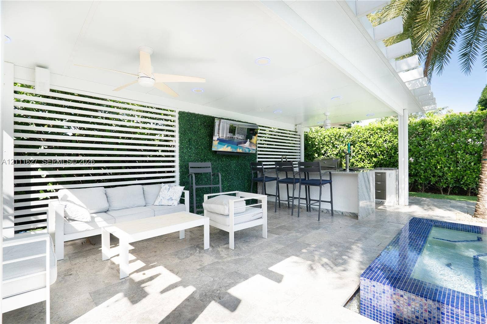 Covered outdoor patio featuring stone flooring, a built-in outdoor kitchen with stone countertops, a slatted privacy screen, integrated ceiling lighting, and a tiled spa