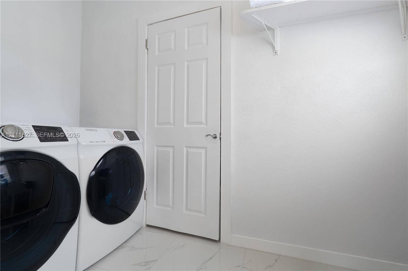 Laundry room featuring large-format tile flooring, a six-panel door with brushed nickel hardware, and an overhead built-in white storage shelf