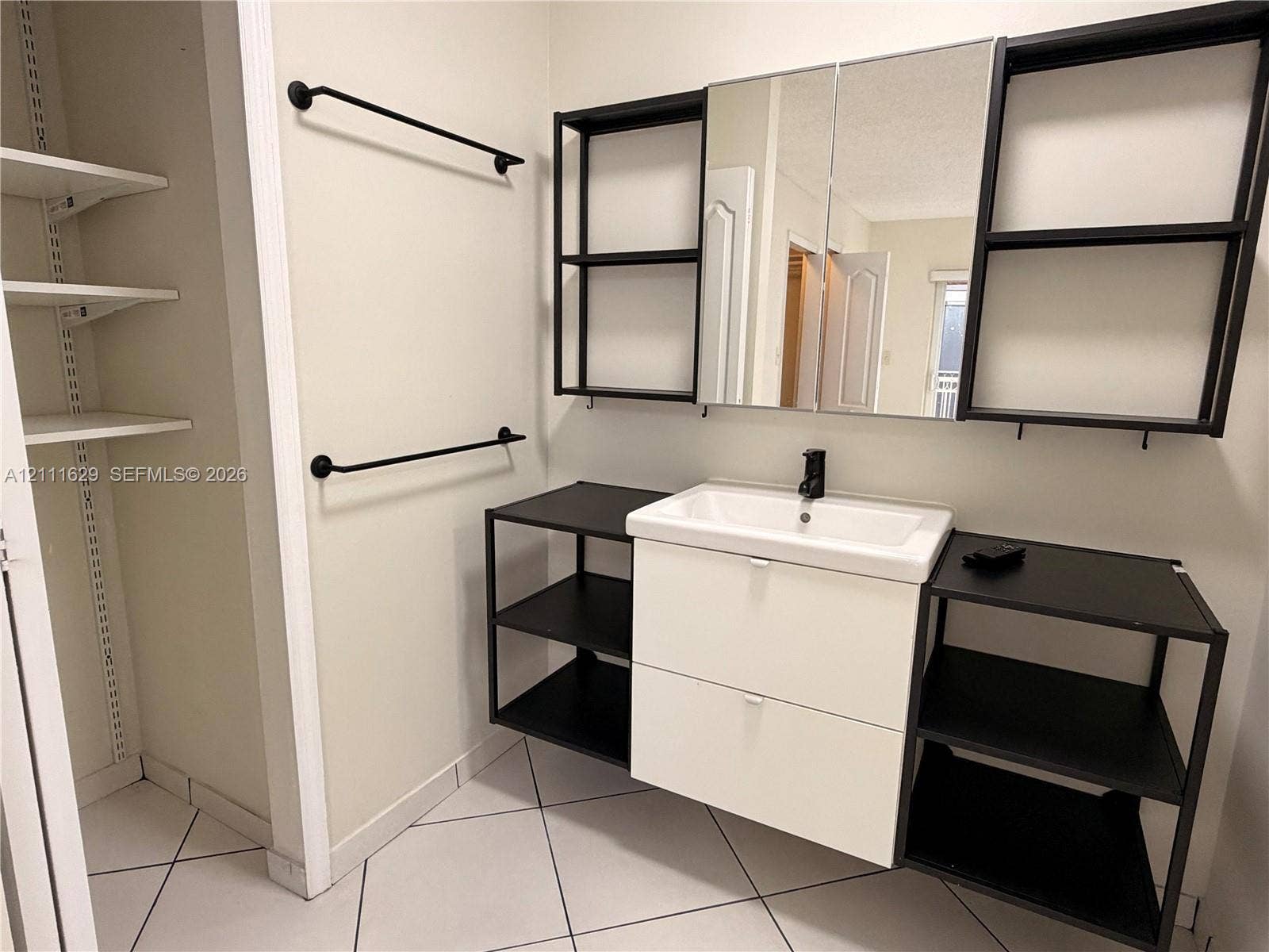 Wall-mounted vanity featuring a rectangular integrated sink, matte black faucet, open metal shelving, and white cabinetry, complemented by a tri-panel mirrored medicine cabinet and tiled flooring