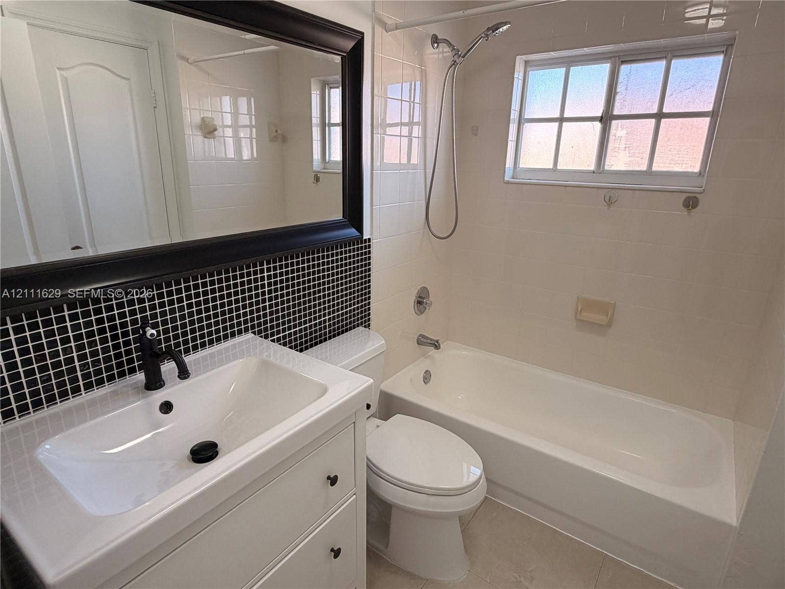 Bathroom featuring a white vanity with a rectangular sink, black faucet hardware, black-and-white grid tile backsplash, framed wall mirror, and a tub-shower combination with a sliding window
