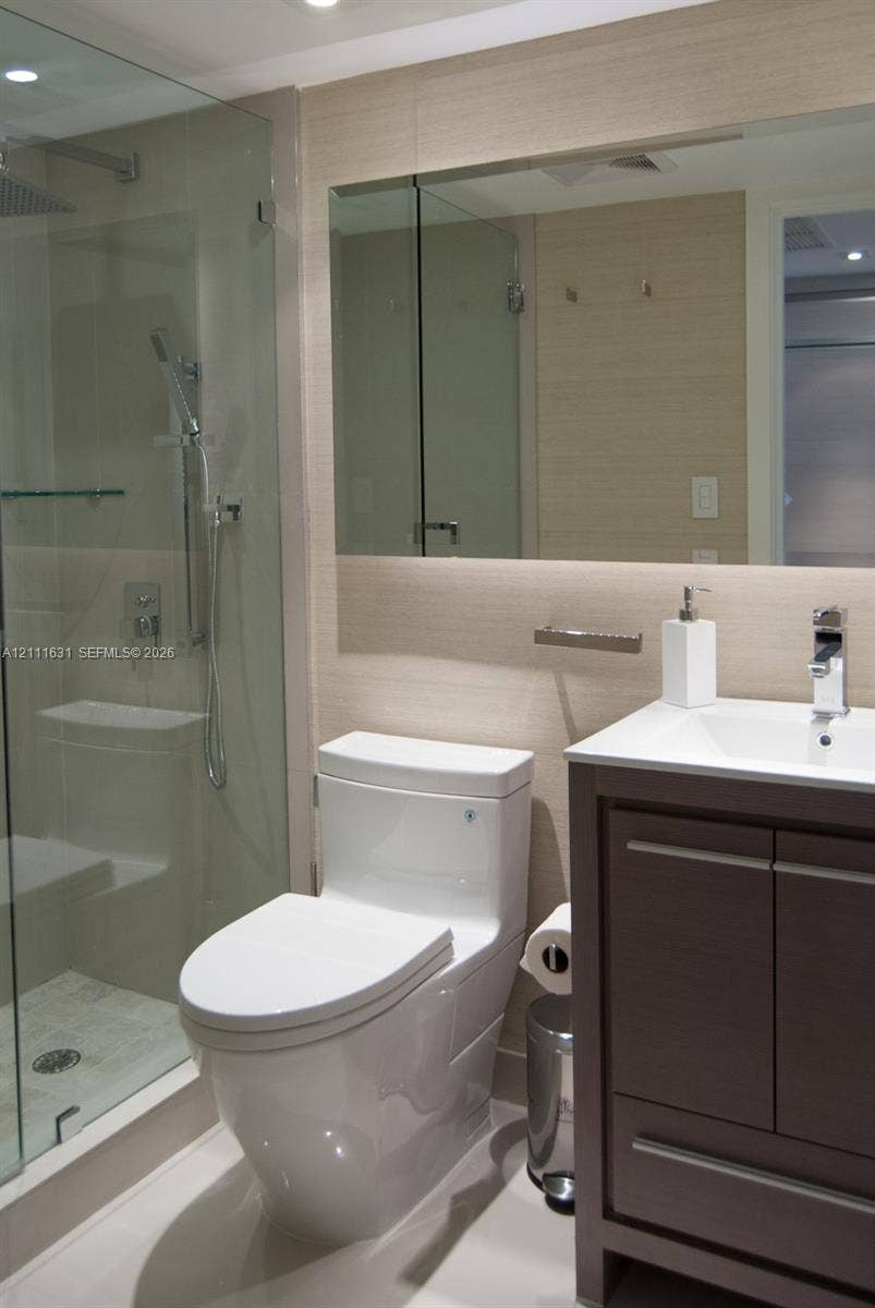 Bathroom featuring a glass-enclosed shower, wood-finish vanity with a rectangular sink, wall-mounted mirror, and neutral wall tile