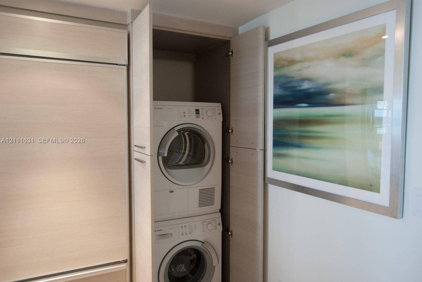 Stacked laundry unit encased in custom cabinetry featuring light-toned wood-finish panels and integrated hardware