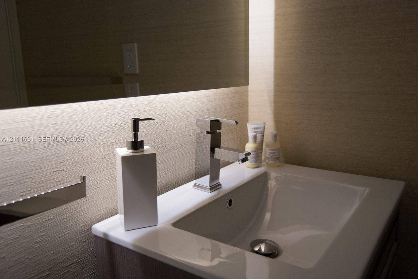 Undermount sink with integrated countertop, polished chrome single-lever faucet, wall-mounted mirror with integrated under-cabinet lighting, and textured wall treatment