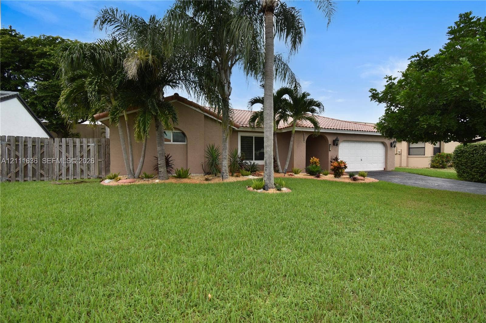 Single-story stucco exterior featuring a tiled roof, multi-car garage, manicured lawn, and mature palm landscaping