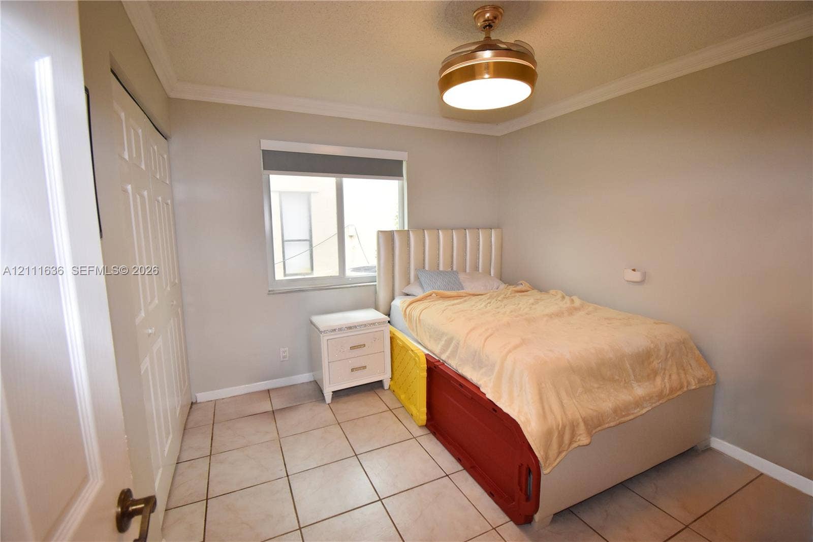 Neutral-toned room featuring tile flooring, crown molding, a dual-panel closet door, a single-hung window with a roller shade, and a flush-mount ceiling fan with integrated light