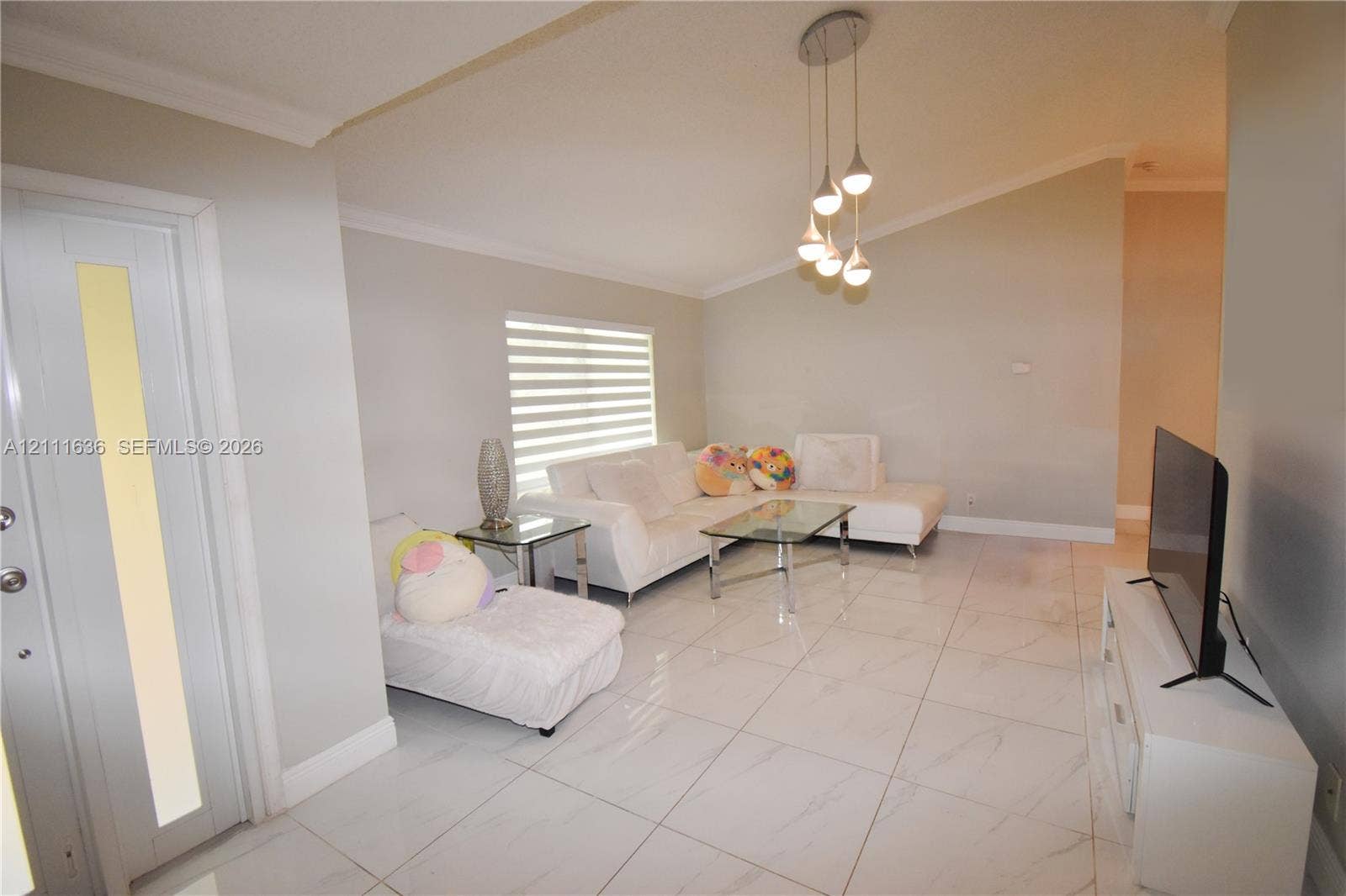 High-ceiling living area featuring large-format polished tile flooring, tiered multi-light pendant fixture, window with horizontal blinds, and glass-paneled entry door