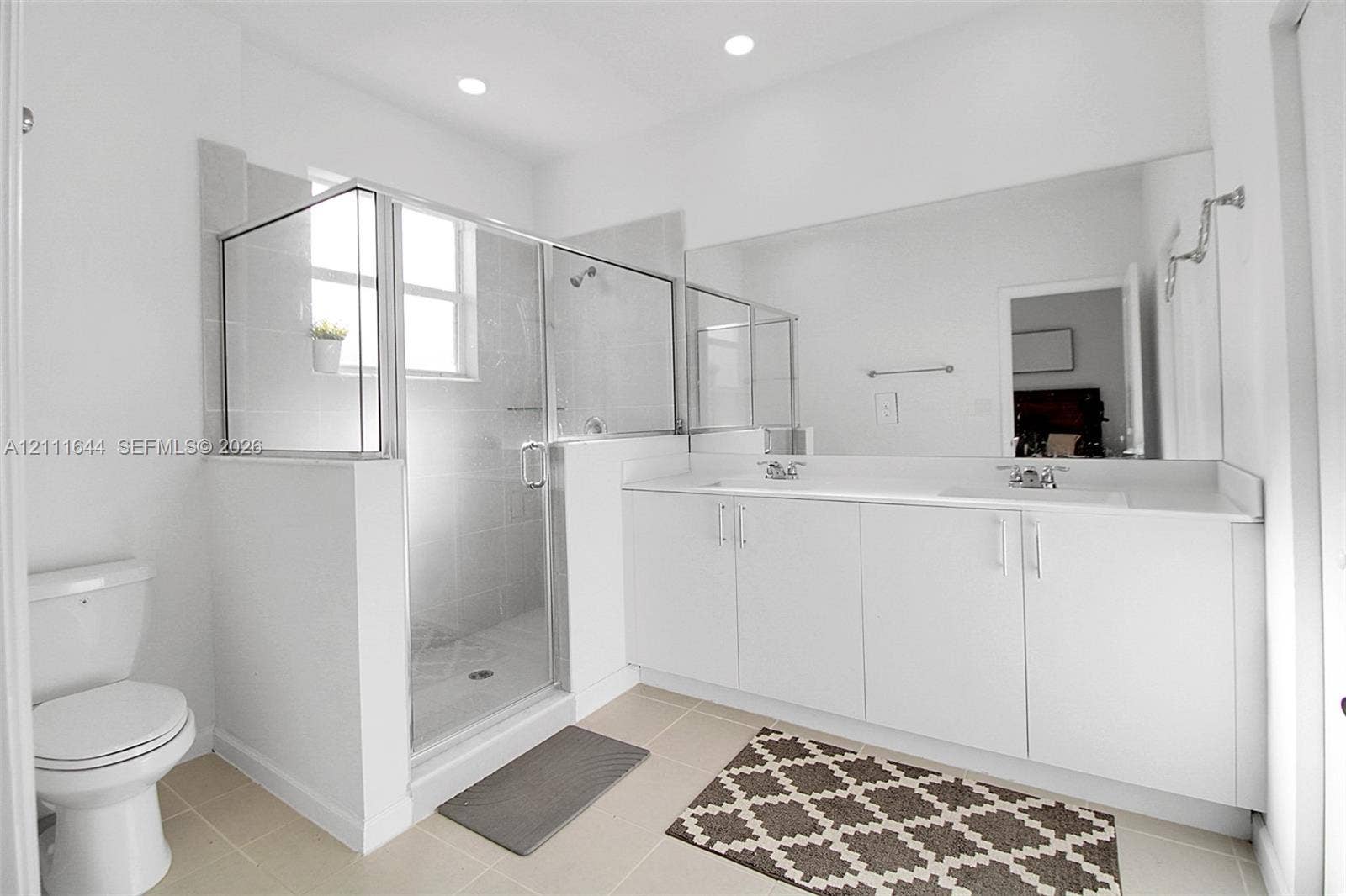 2873 SE 24th Avenue, Homestead, FL 33035 Double vanity with white cabinetry and solid surface countertop, large wall-mounted mirror, and glass-enclosed shower with tiled surround