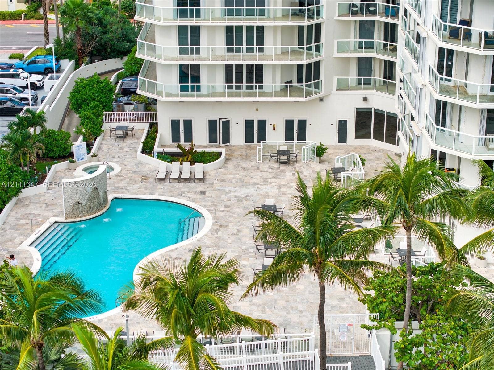 8925 Collins Avenue, #4J, Surfside, FL 33154 Multi-story building featuring tiered balconies with glass railings, expansive stone-paved pool deck, swimming pool with integrated spa and water feature, and established tropical landscaping