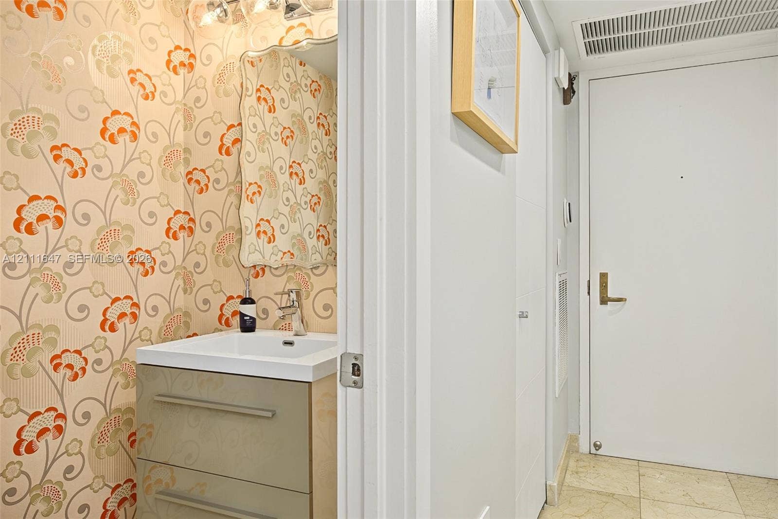 8925 Collins Avenue, #4J, Surfside, FL 33154 Powder room featuring floral patterned wallcovering, wall-mounted mirror, two-drawer vanity with a white basin, and polished metal hardware