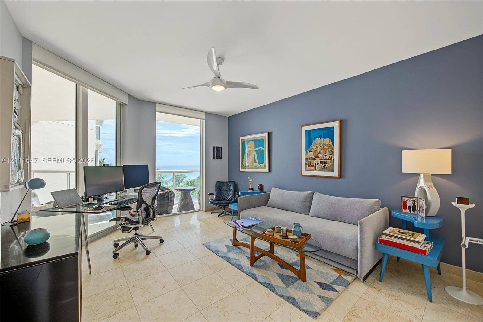 8925 Collins Avenue, #4J, Surfside, FL 33154 Floor-to-ceiling sliding glass doors, light-toned stone tile flooring, neutral wall palette with a prominent blue accent wall, ceiling fan with integrated lighting, and balcony access