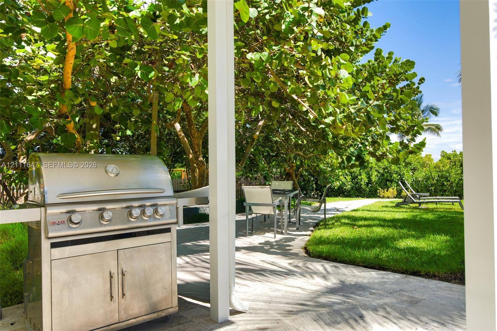 8925 Collins Avenue, #4J, Surfside, FL 33154 Covered patio featuring a built-in stainless steel grill, stone paver flooring, and mature landscaping surrounding a grass lawn