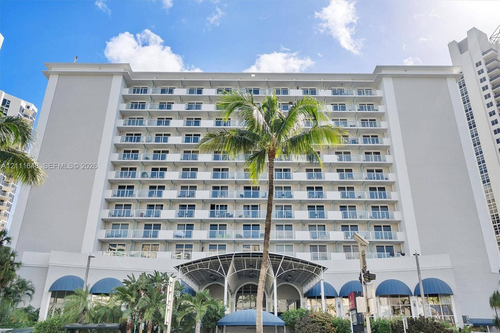 19201 Collins Avenue, #518, Sunny Isles Beach, FL 33160 Mid-rise building exterior featuring multiple levels of private balconies with glass railings, a prominent arched entrance canopy, and blue architectural awnings