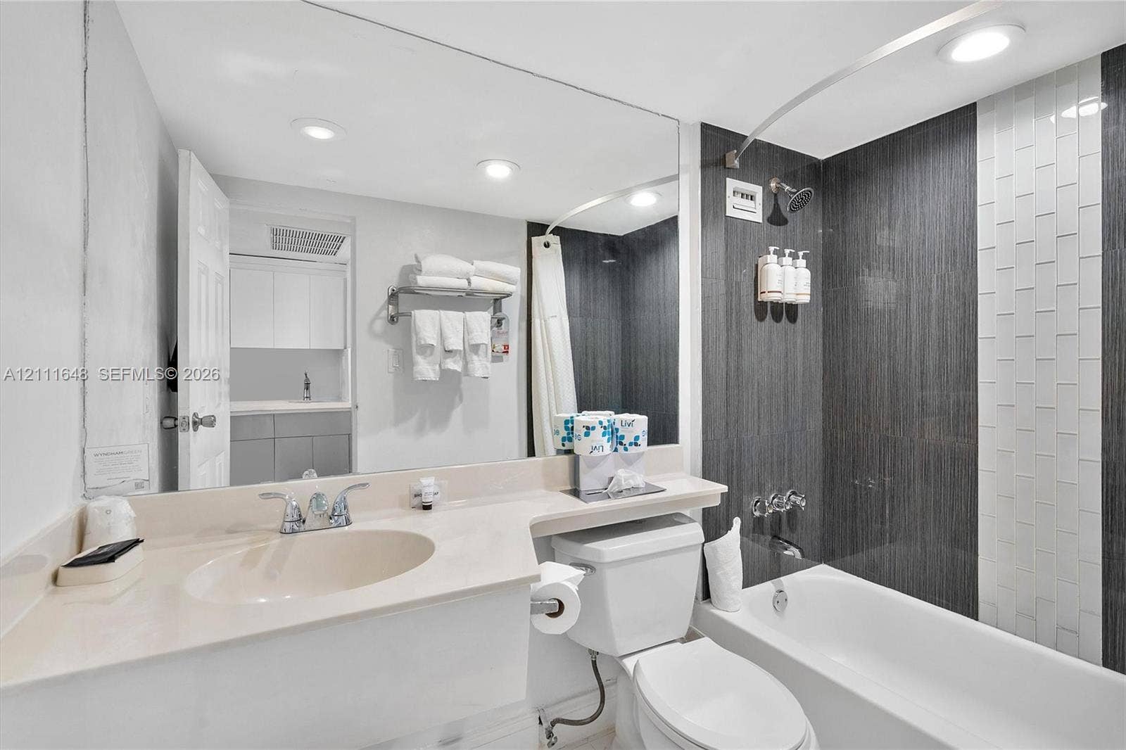 19201 Collins Avenue, #518, Sunny Isles Beach, FL 33160 Full-width vanity with an integrated sink, expansive wall-mounted mirror, recessed lighting, and shower area featuring vertical charcoal and white tile surround