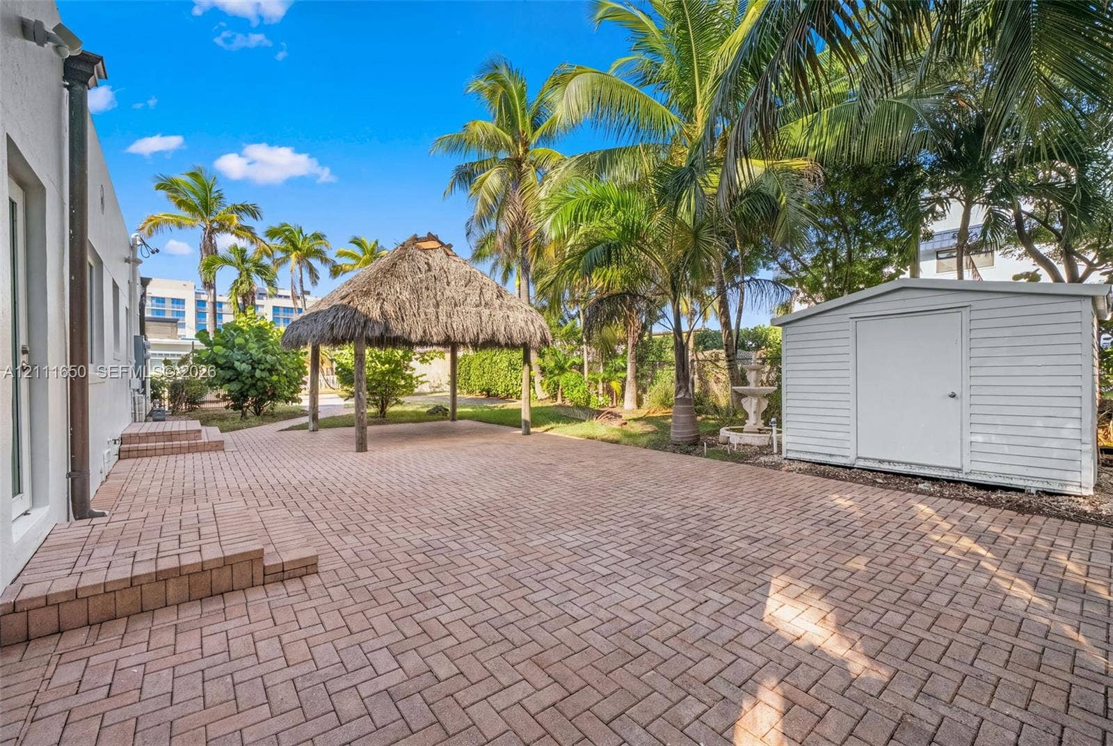 Expansive brick-paver patio featuring a thatched-roof tiki hut, white vinyl storage structure, decorative stone fountain, and mature palm trees